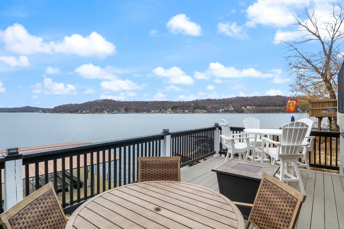 The image showcases a serene lake view from the deck, featuring outdoor seating and a table set up for dining or relaxing. The tranquil water, framed by trees, creates an idyllic setting for people and pets to unwind together and enjoy nature.
