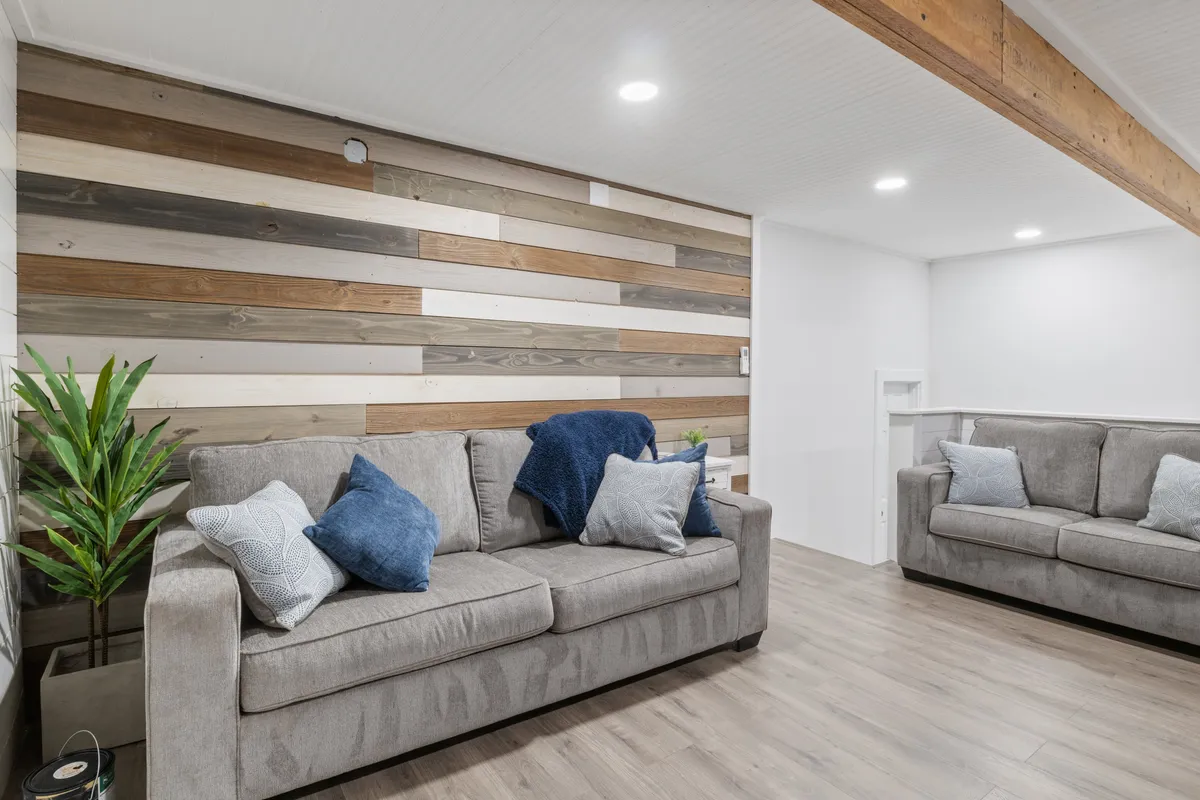 A cozy living room corner with sofas, a wooden accent wall, decorative pillows, and an indoor plant. The arrangement encourages socializing, game nights, or quiet relaxation.