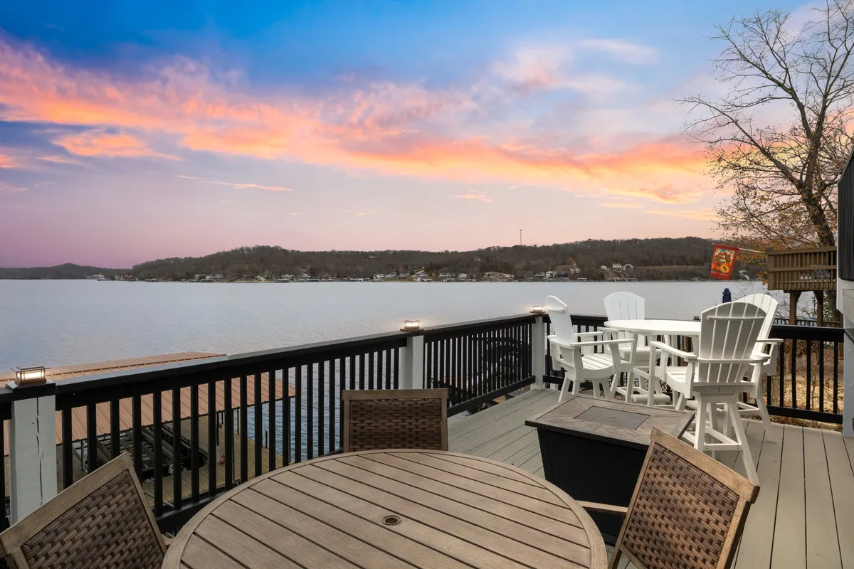 The image shows an expansive lake view at sunset, with vibrant colors painting the sky. In the foreground, comfortable outdoor furniture invites relaxation and socializing on the two-tier deck. The setting is tranquil and designed for outdoor dining, barbecues, and soaking in peaceful lakeside moments.