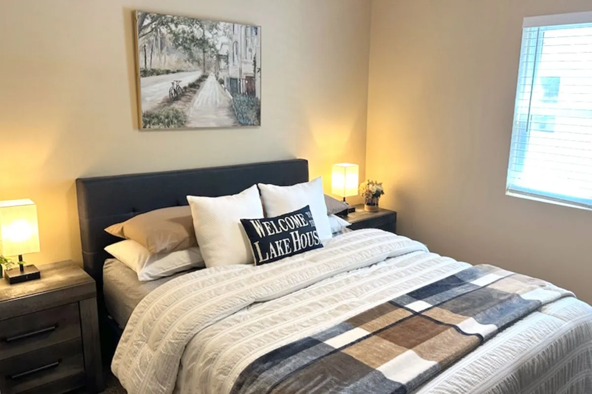 A cozy, well-lit bedroom featuring a plush queen bed adorned with decorative pillows, nightstands with lamps on either side, and a tasteful painting above the headboard. Soft natural light filters through the window, creating a restful and inviting atmosphere.
