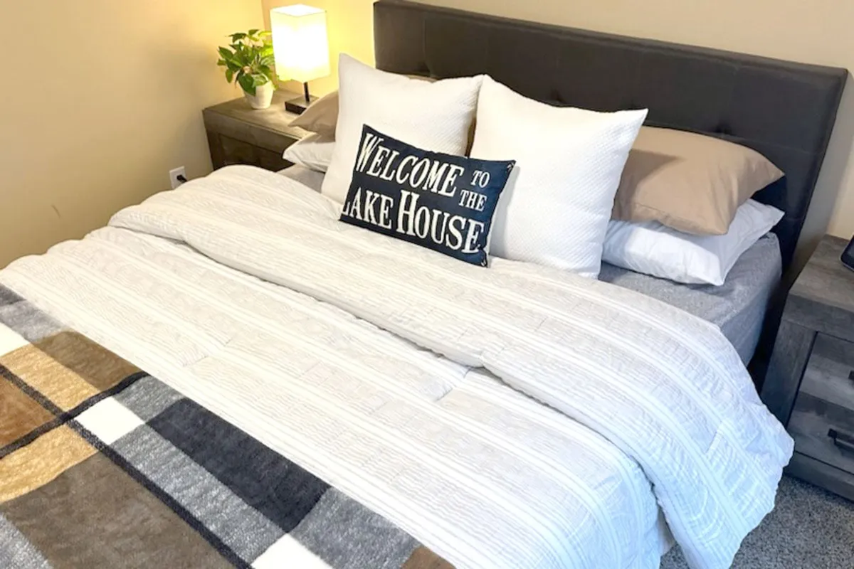 Another serene bedroom scene, showing a neatly made bed with decorative pillows, a nightstand with a lamp, and a plaid throw blanket. The room exudes warmth and comfort, encouraging relaxation and cozy evenings.