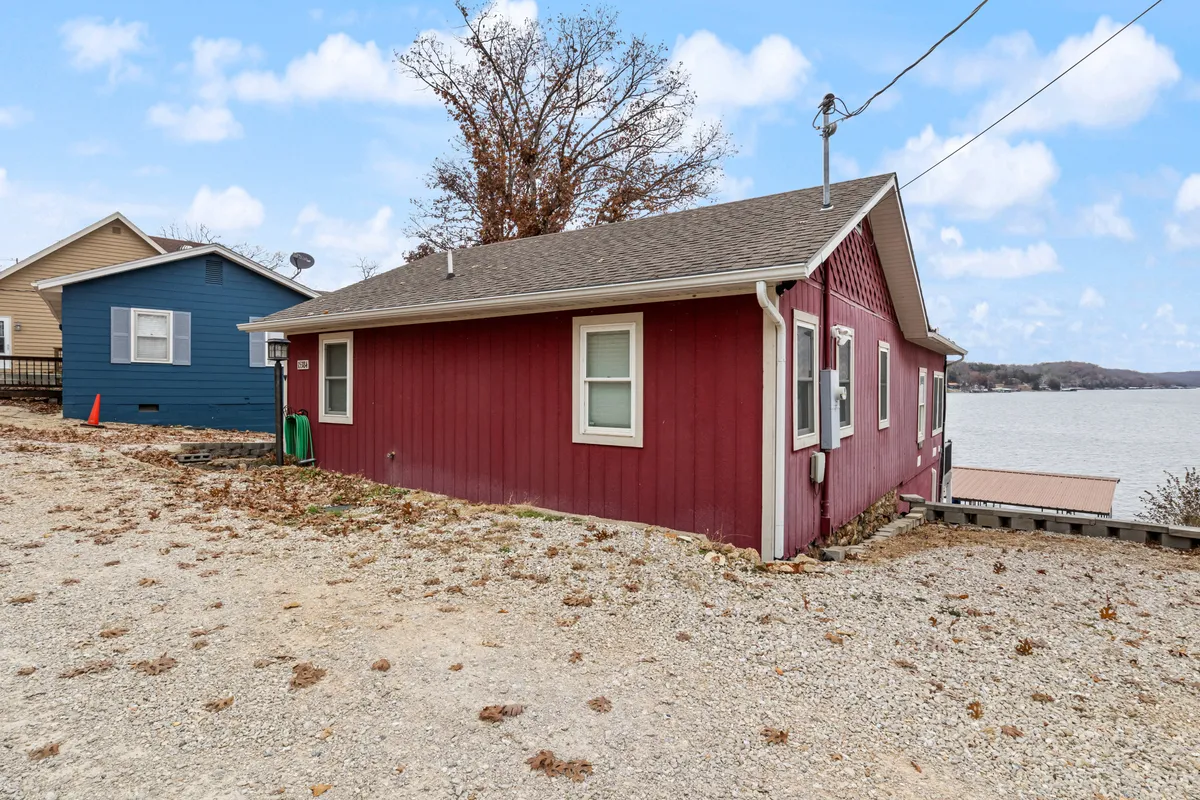 This exterior shot captures the red wooden house with a beautiful lake view and a neighboring blue house, set in a casual, serene environment. The scene hints at direct access to the outdoors and proximity to nature trails.