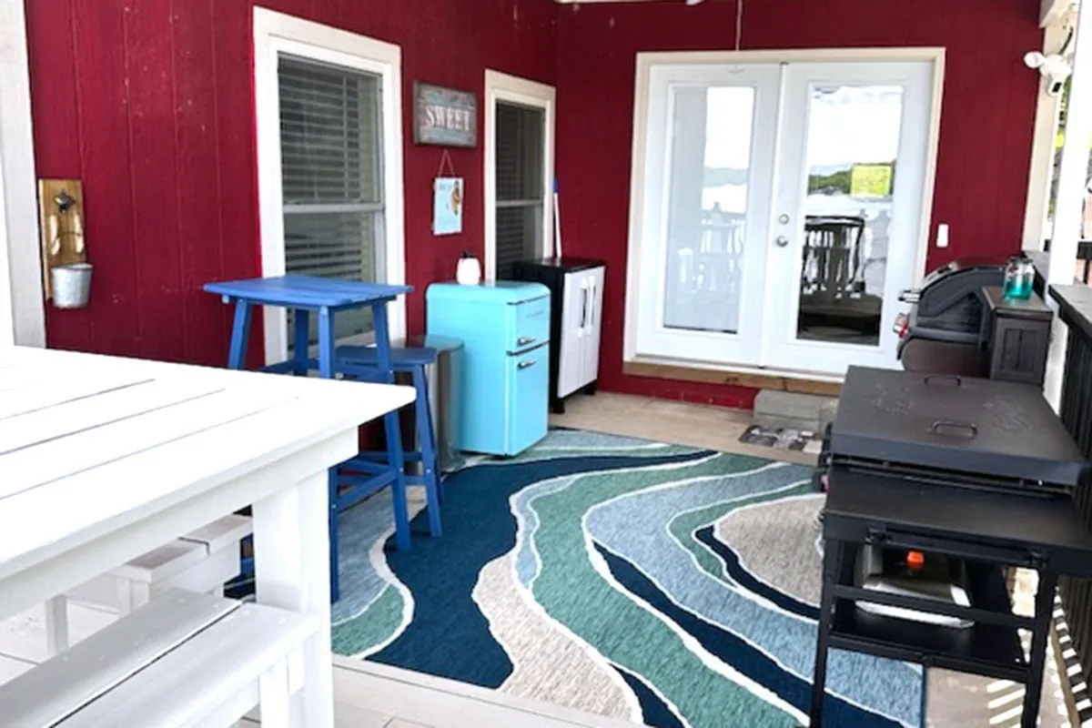 A vibrant deck scene with red walls, blue furniture, a decorative rug, and white doors. Friends relax and dine outdoors as the sun sets, with the BBQ grill in use and plenty of space for conversation and games.