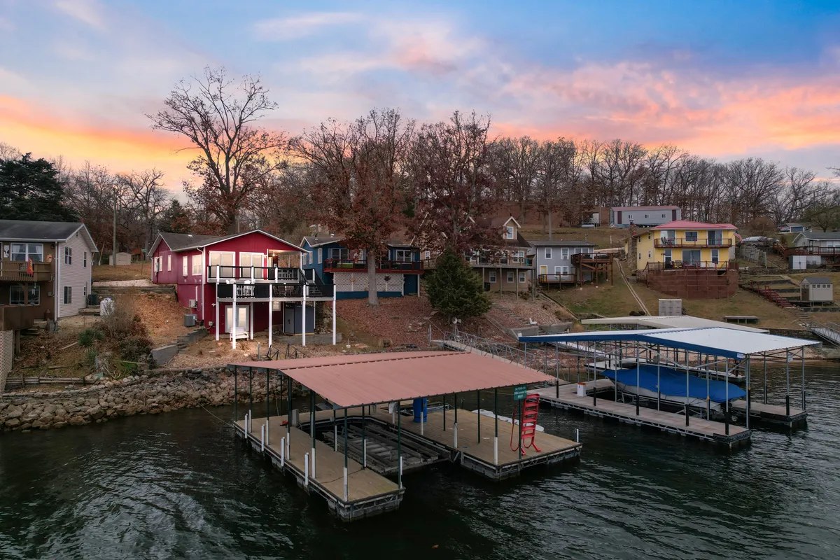 A panoramic lakeside view with colorful cottages and docks set along the tranquil water’s edge, framed by lush trees. Several boats are visible along the docks, while people enjoy water-based activities and dockside relaxation.