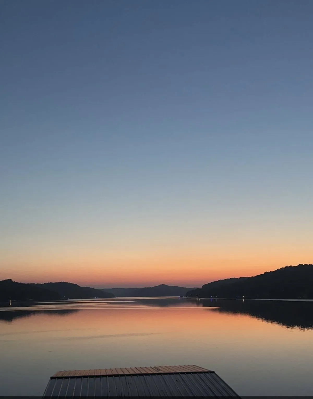 A tranquil lakeside scene at sunset, where soft colors paint the sky over the horizon and distant hills. The calm water invites activities like boating and fishing, while the peaceful ambiance hints at stargazing or sharing meals by the water's edge.