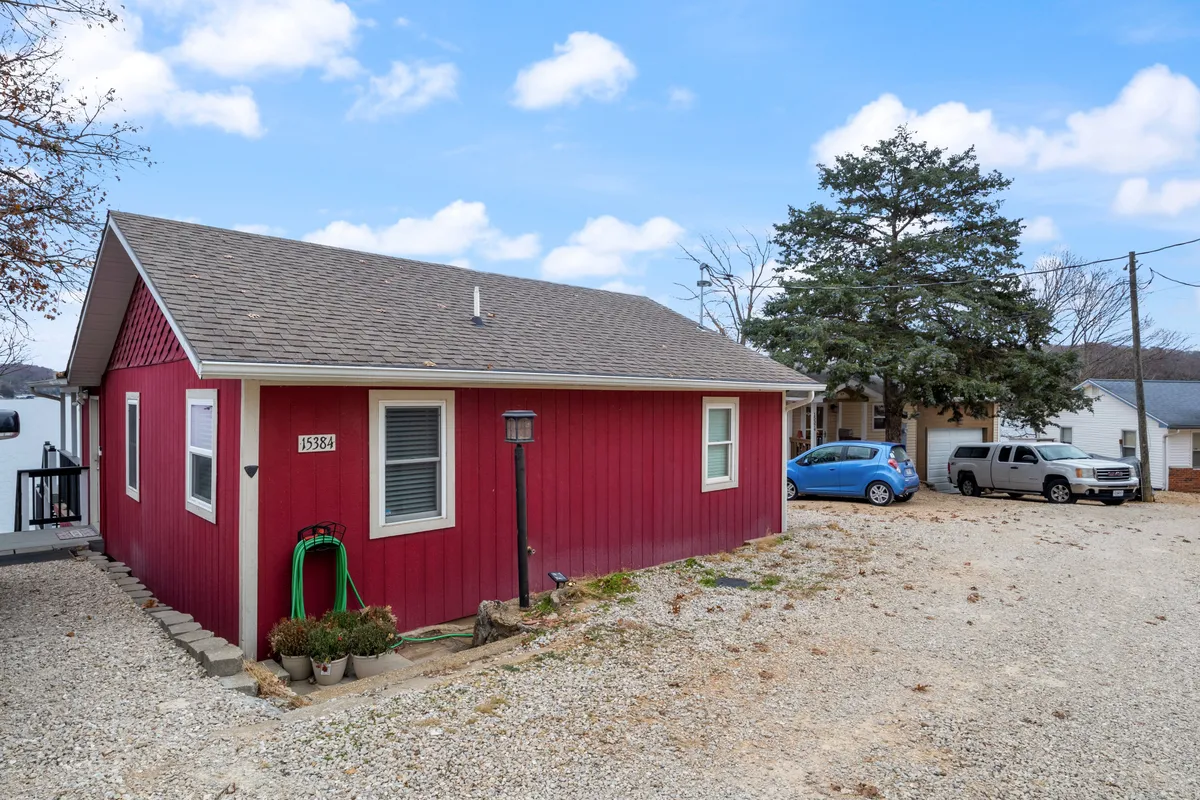 This exterior image features the red facade of the Melo Escape house, a welcoming lamp post, a gravel driveway, and lush trees surrounding the property. The setting evokes a homey, inviting atmosphere with space for outdoor gatherings and easy access to nature.
