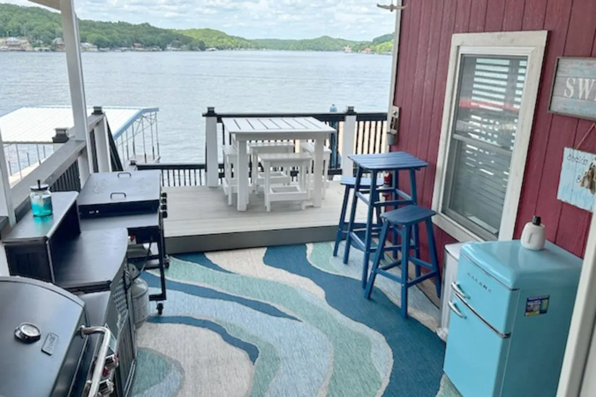 An inviting outdoor dining area on the deck, featuring a large table surrounded by blue stools, a grill, vintage refrigerator, and a decorative rug. The setting is cheerful and laid-back, perfect for gatherings and al fresco meals.