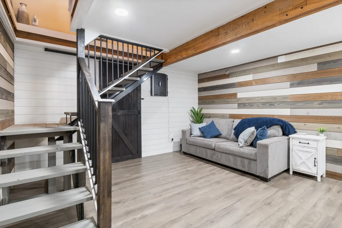 A living area with a grey pullout couch accented by blue pillows, set against a textured wood wall and adjacent to a staircase. There is ample natural light, and the space invites relaxation or socializing, making it ideal for flexible sleeping arrangements.